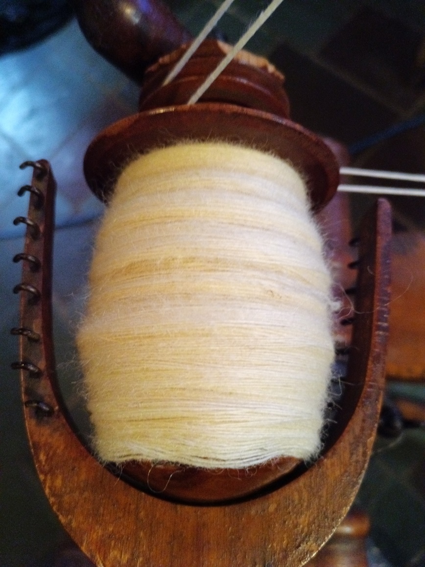 A wooden wheel bobbin full of shiny white singles yarn that has a halo.