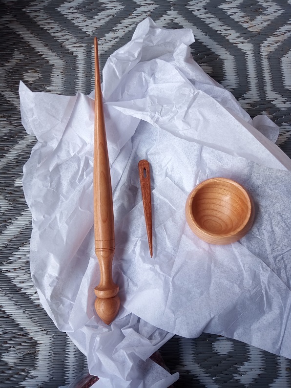 A smooth brown supported spindle made of wood is on the left, a small brown nalbinding needle is next to it and a tan supported spindle bowl on the right.
