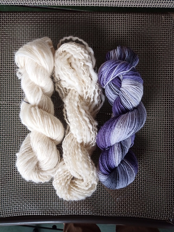 Three skeins of yarn on a mesh grey chair. From left to right they are white singles puffy yarn, white spiral ply yarn where the thicker ply puffs around the thinner ply, and purple to grey gradient yarn that is smooth.