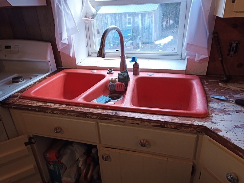 A deep orange three bay sink is centered in the photo. The counter around it is brown and mottled but free of tile.