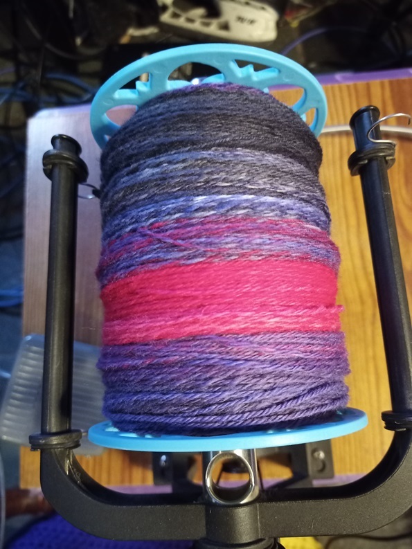 A bobbin of soft, squishy yarn that is black, grey, purple and red.